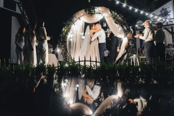Para Młoda podczas romantycznej ceremonii ślubnej w plenerze, udekorowanej okrągłym łukiem z gałęzi i światełkami – modne trendy weselne 2026